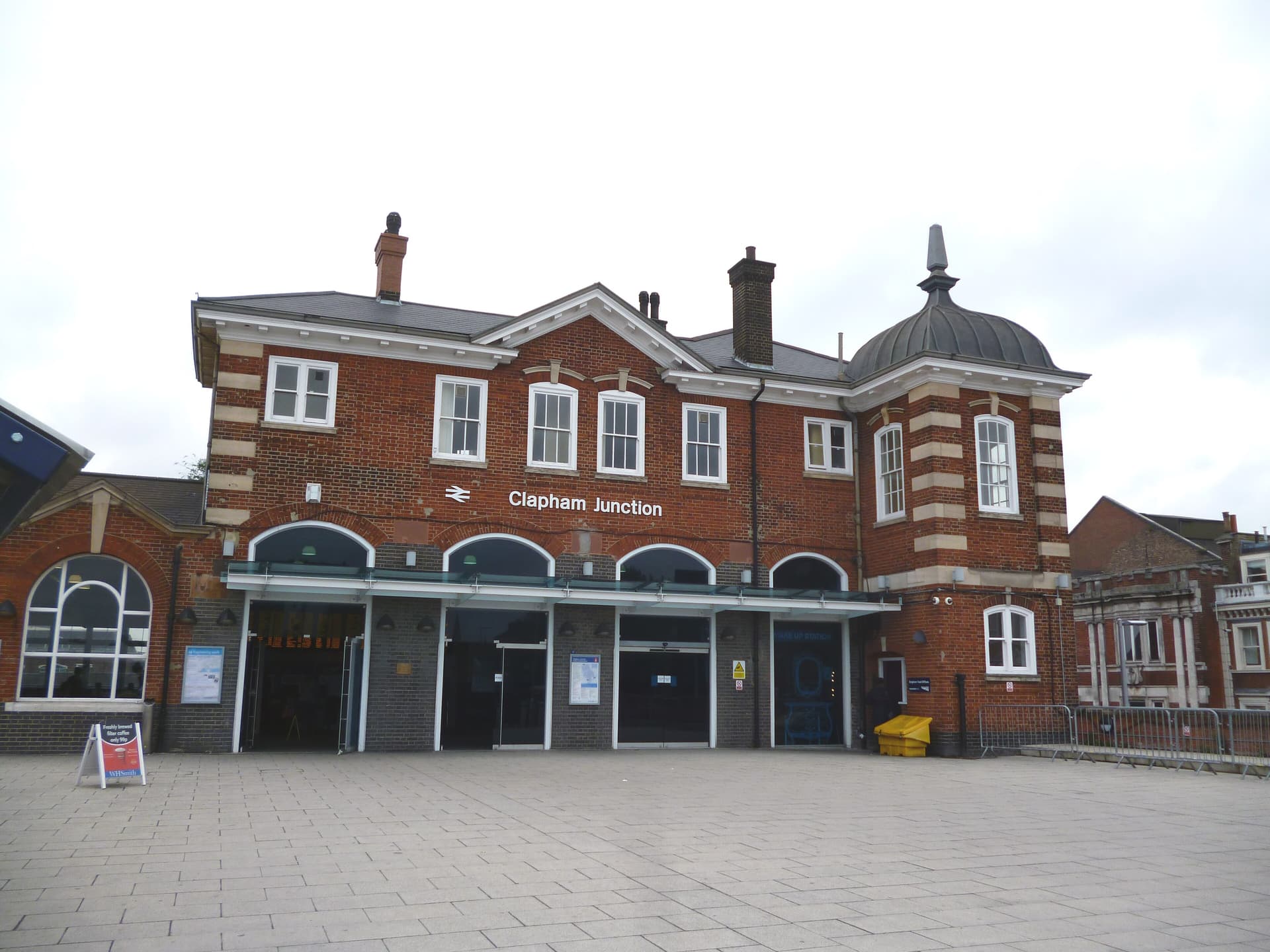 Red-brick Edwardian frontage of Clapham Junction station with arched entrances, a domed corner turret and an empty paved forecourt
