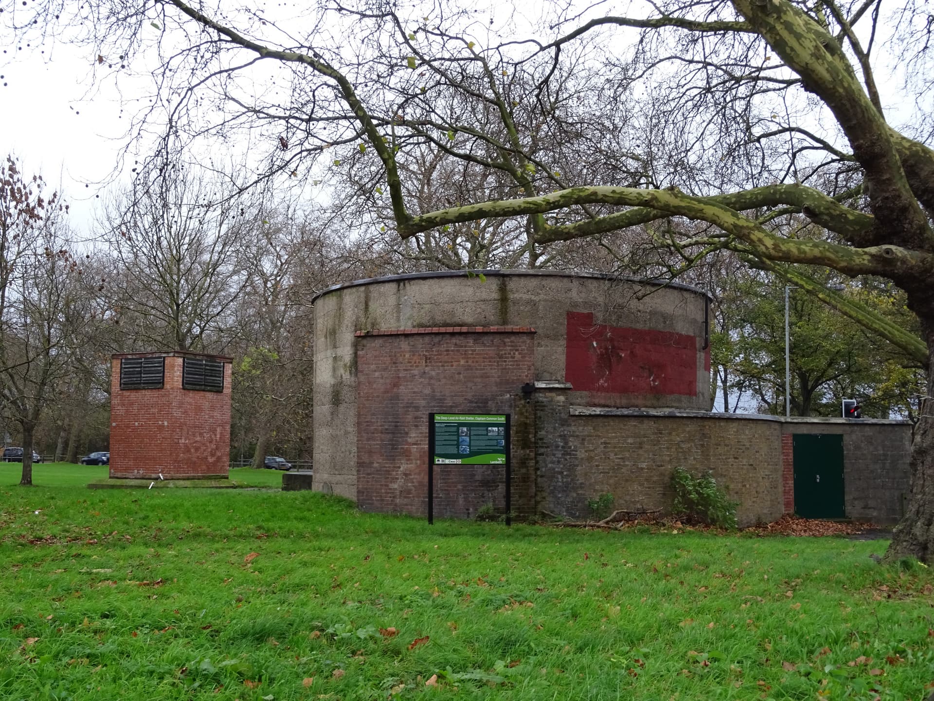 Circular concrete and brick deep-level air-raid shelter entrance beside a brick ventilation shaft on grass, Clapham Common