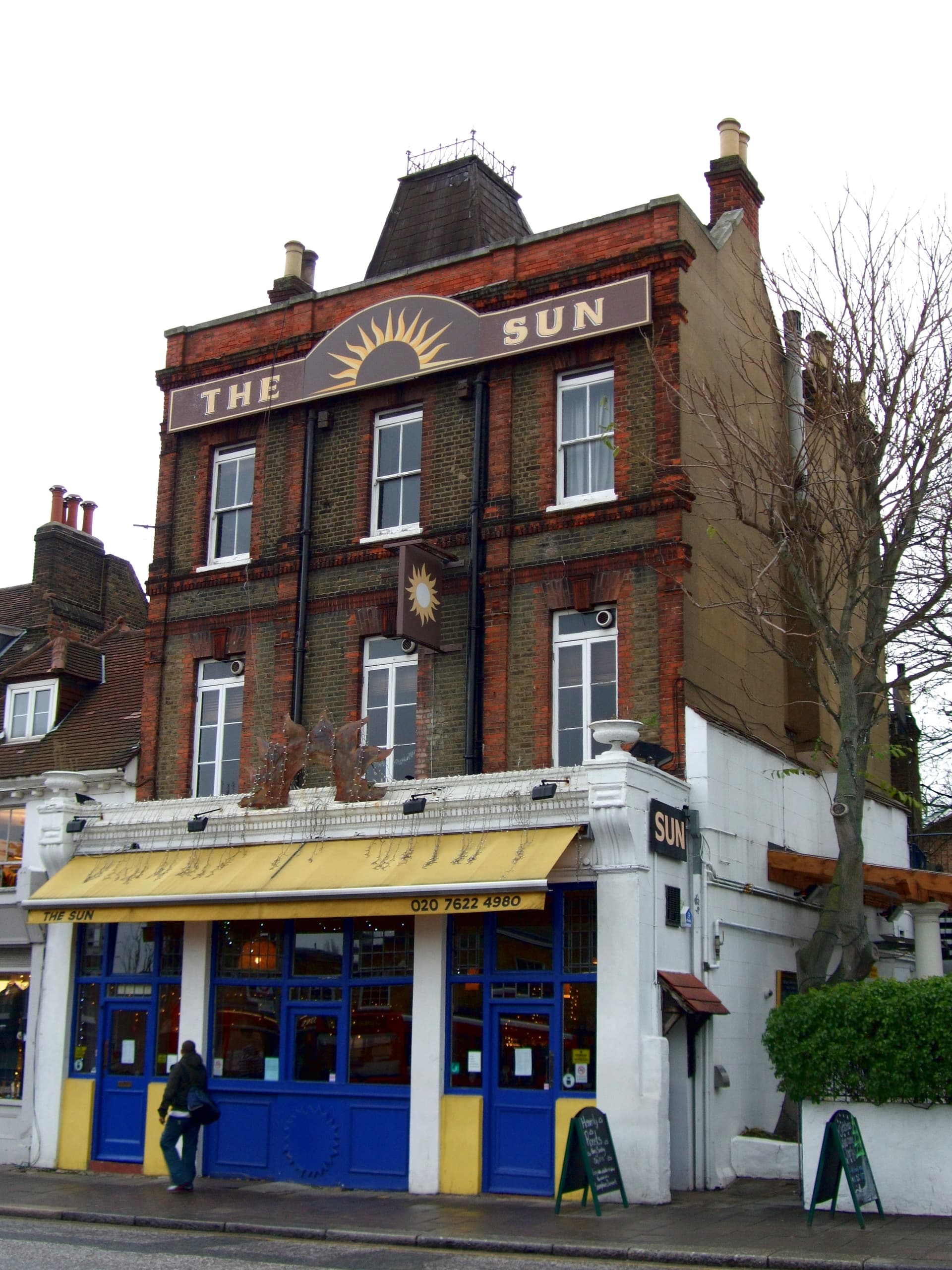Three-storey Victorian brick pub with a sun emblem, yellow awning and blue shopfront on a London street