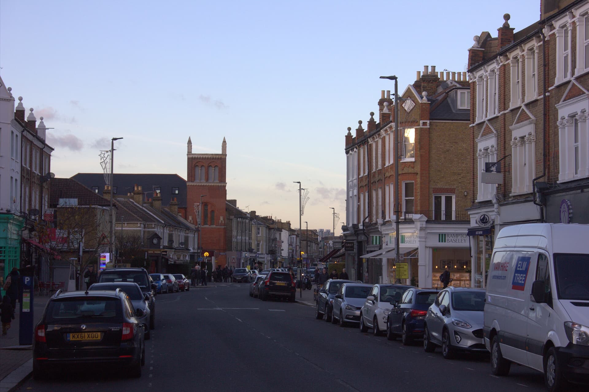 Dusk view along a Battersea shopping street lined with Victorian brick buildings, parked cars and a red church tower beyond