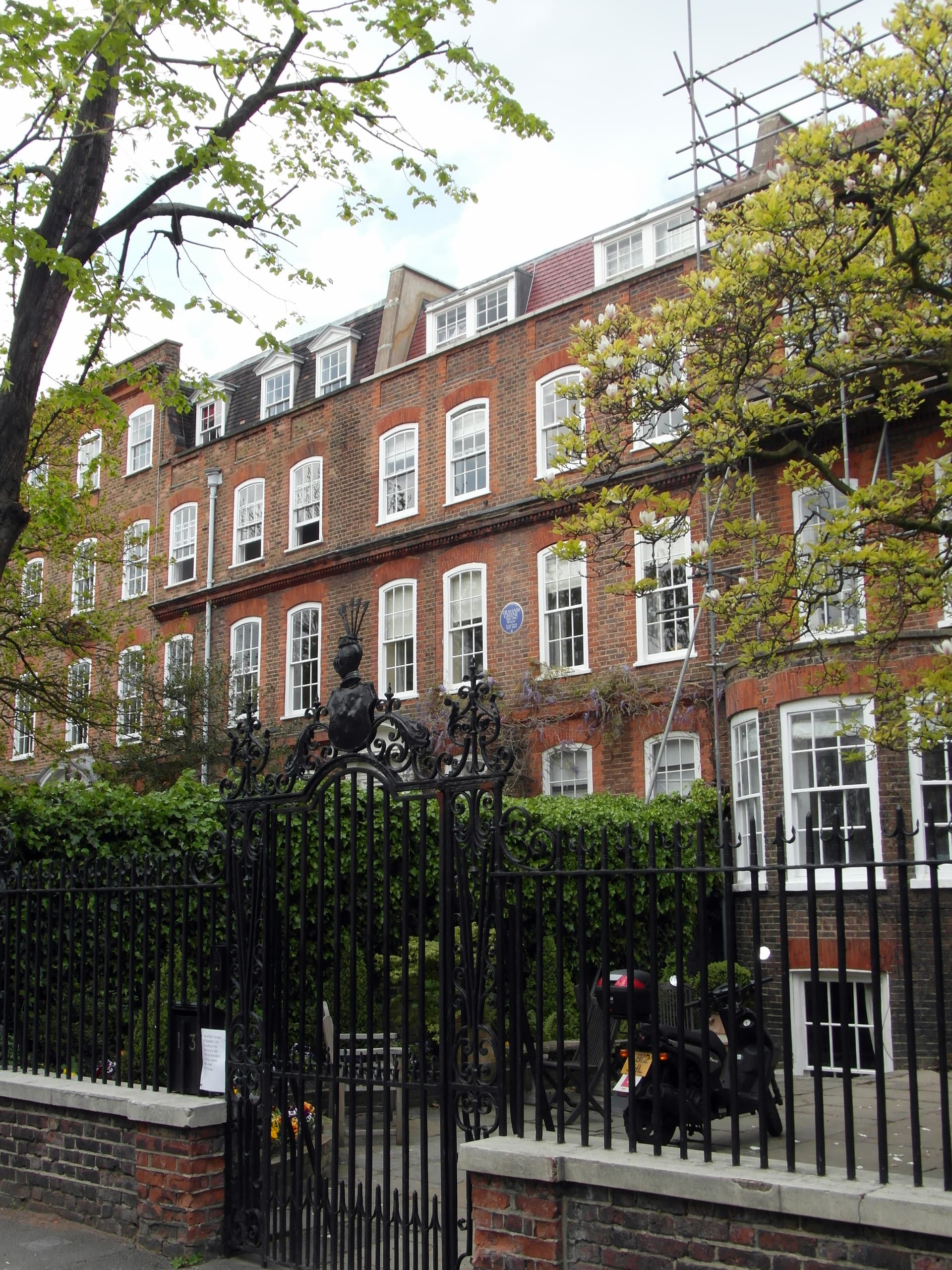 Red-brick Georgian terrace behind black wrought-iron railings and gate, with magnolia blossom and scaffolding on the roof