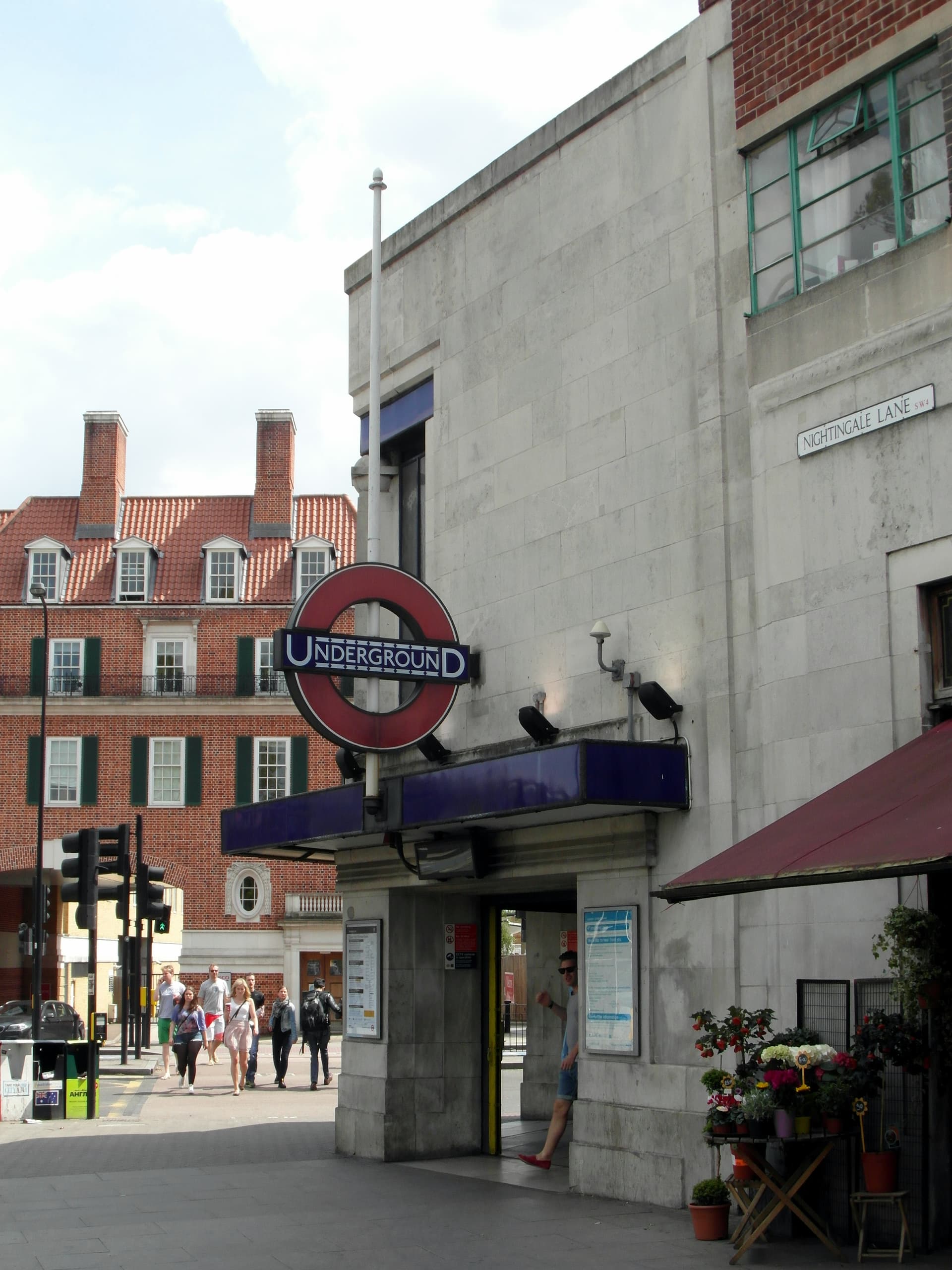 Stone-fronted Underground station entrance with roundel, flower stall and red brick houses beyond, on Nightingale Lane corner in south London