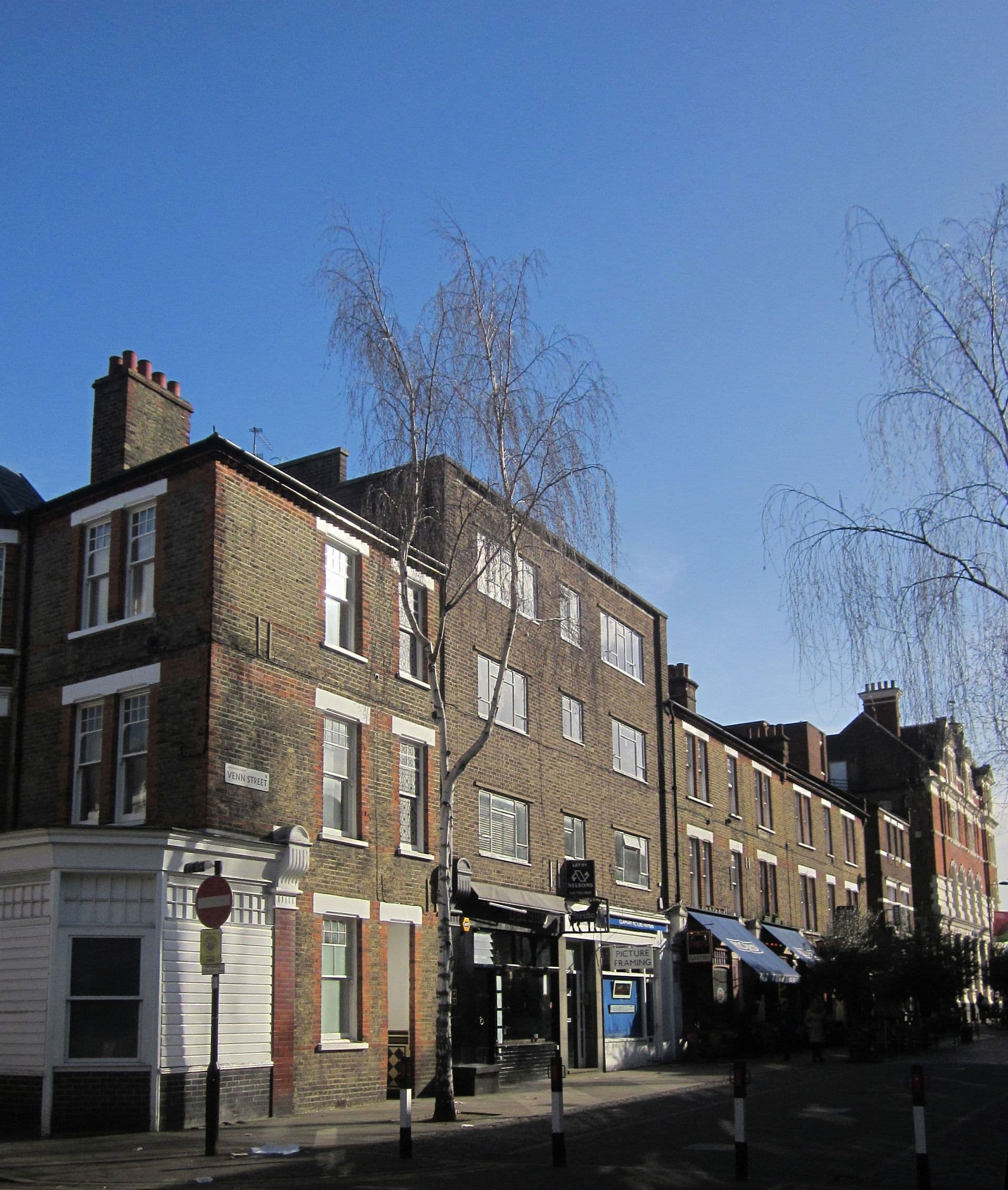 Brick terraced buildings on Venn Street, Clapham, with birch trees, shopfronts and awnings under a clear blue sky