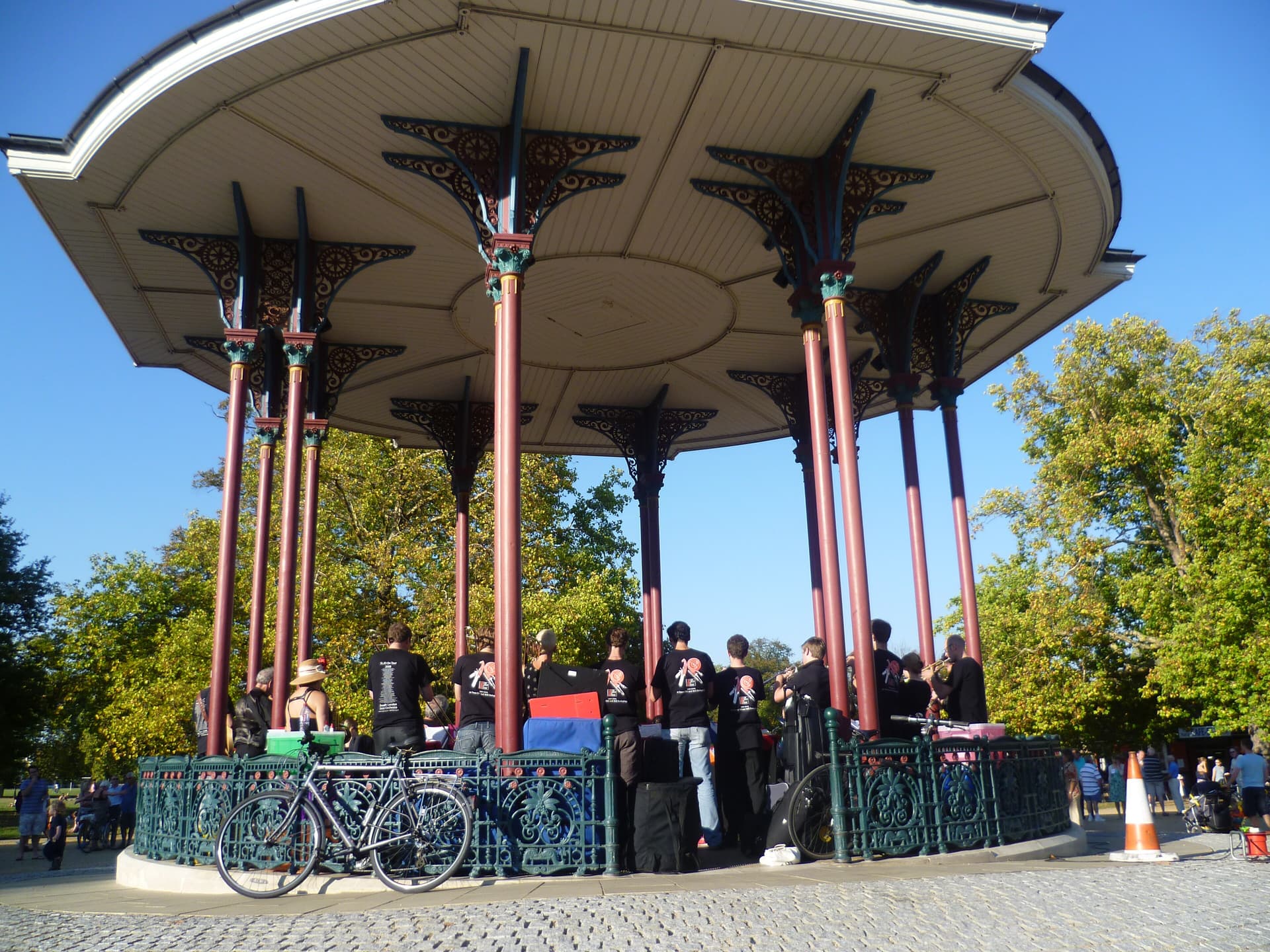 Band in black T-shirts playing under an ornate Victorian bandstand on a sunny day, with trees and a bicycle