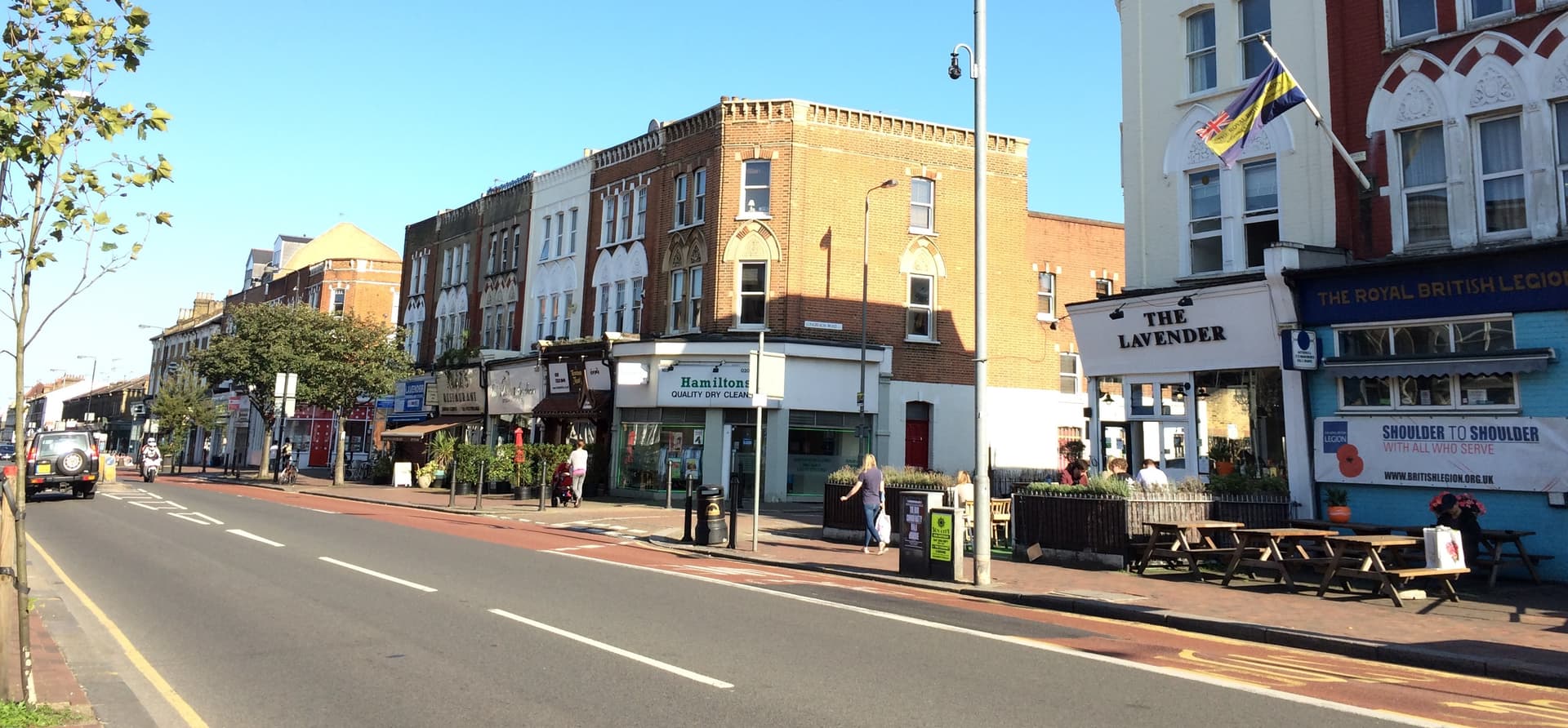 Sunlit Victorian shopfronts and a pub terrace along a London high street with a red bus lane