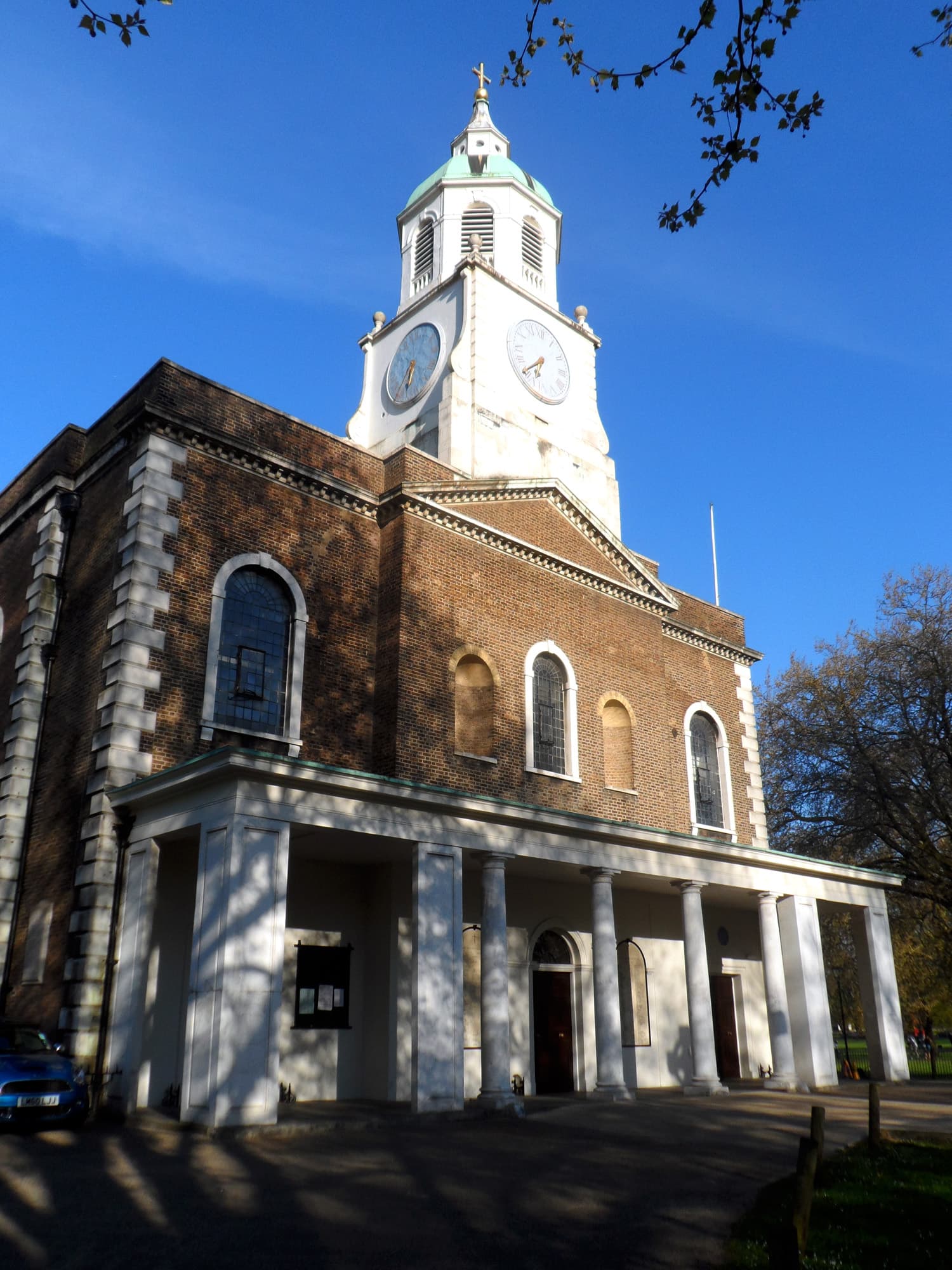Brick Georgian church with white clock tower, copper dome and columned portico beside trees under a clear blue sky
