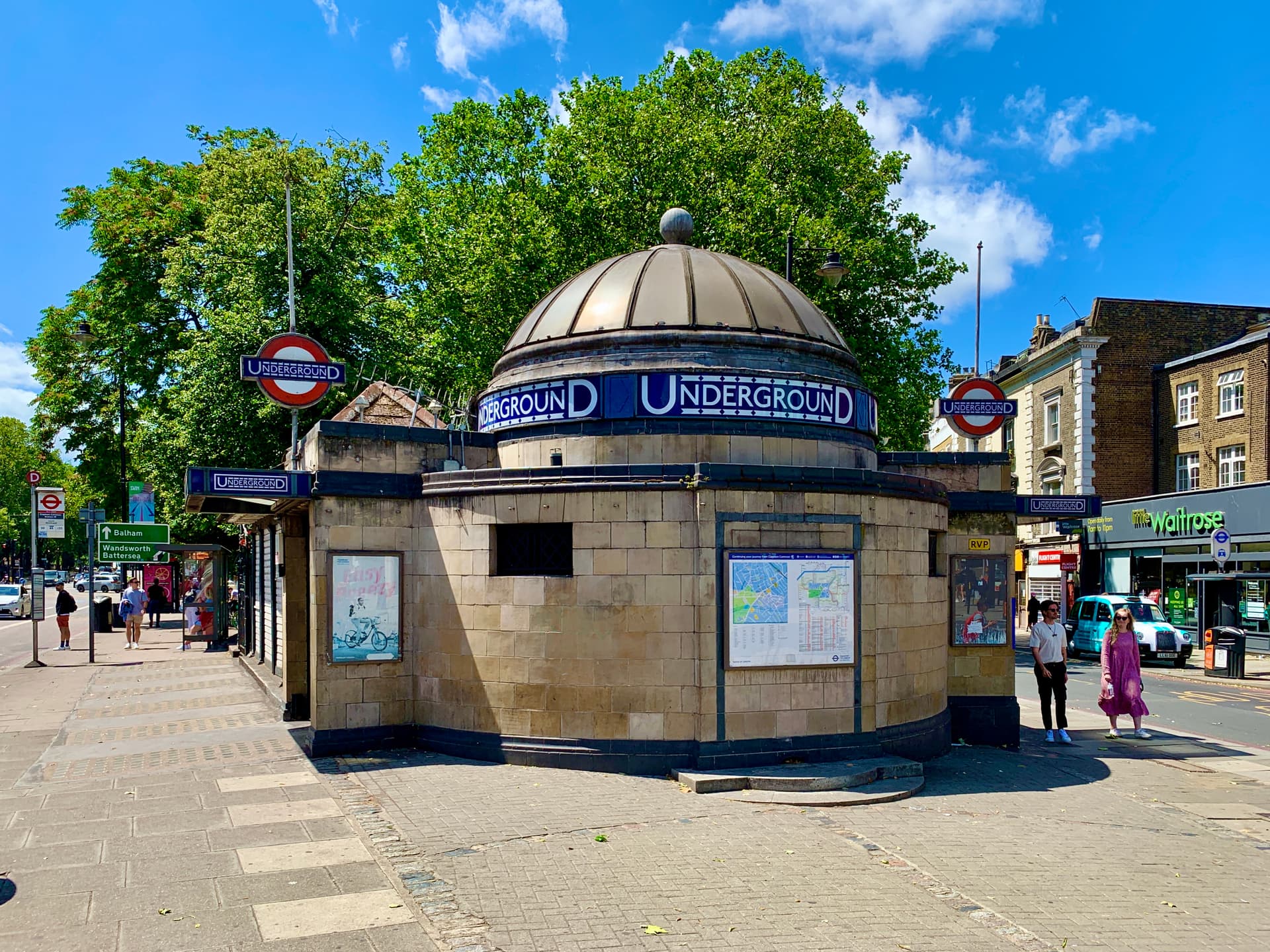 Domed tiled Underground station entrance at Clapham Common on a sunny day, with trees, bus stop and shops behind