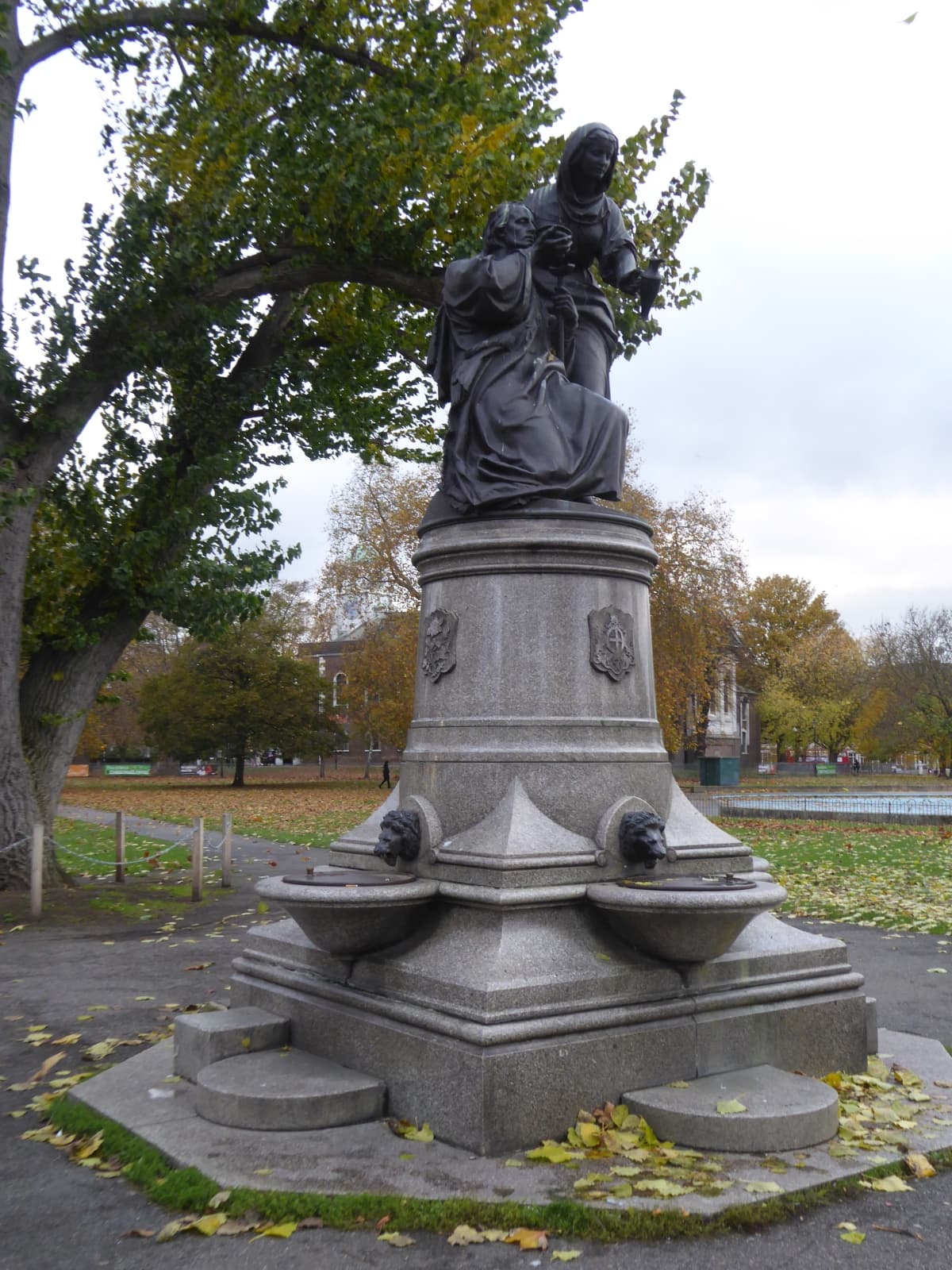 Bronze statue of two figures on a granite fountain base with lion heads, beside autumn trees on the common