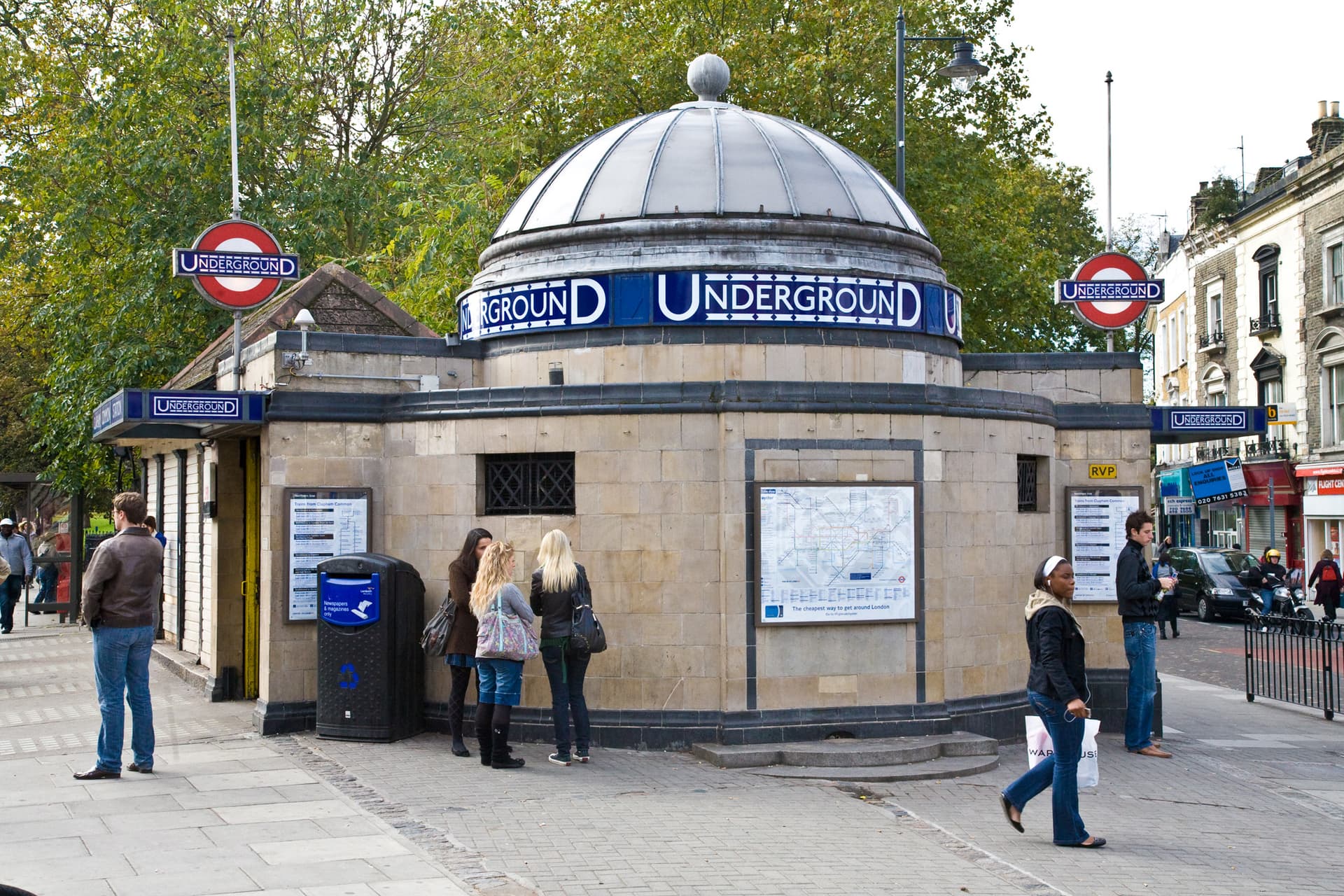 Domed tiled Underground station entrance with roundels, surrounded by trees, with pedestrians on the pavement outside