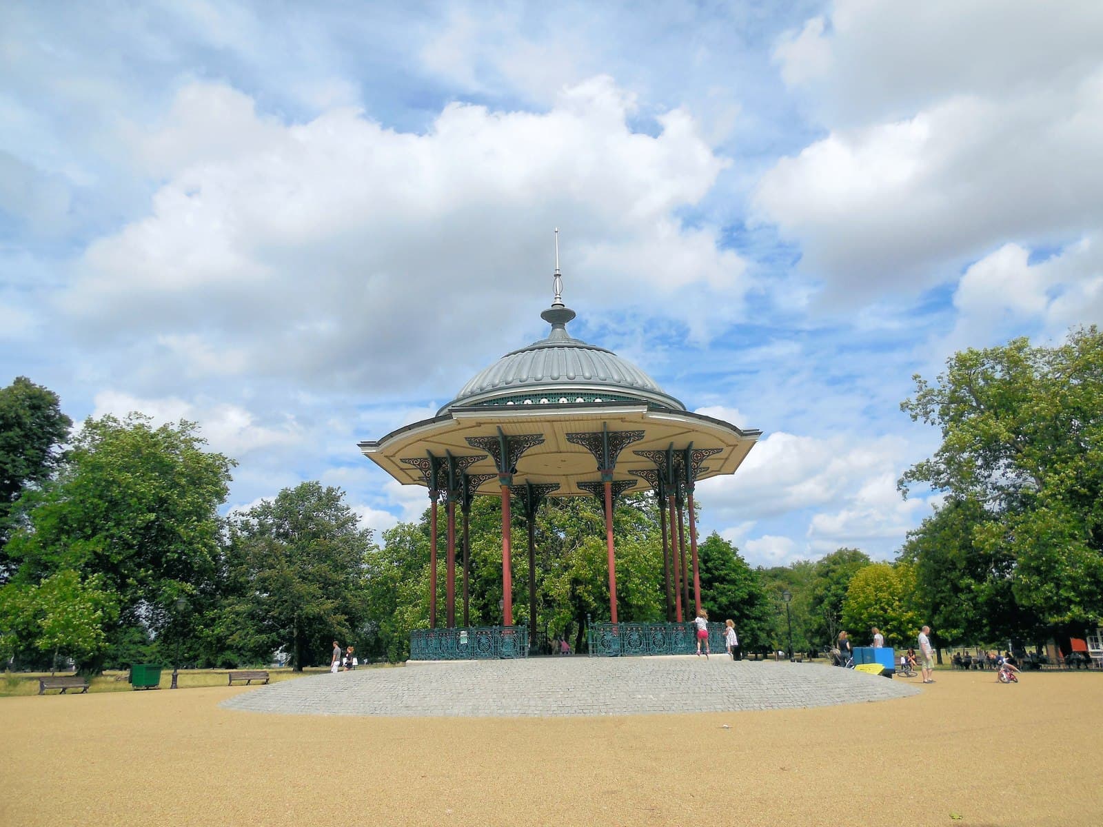 Victorian cast-iron bandstand with a domed roof on Clapham Common, surrounded by trees under a cloudy blue sky
