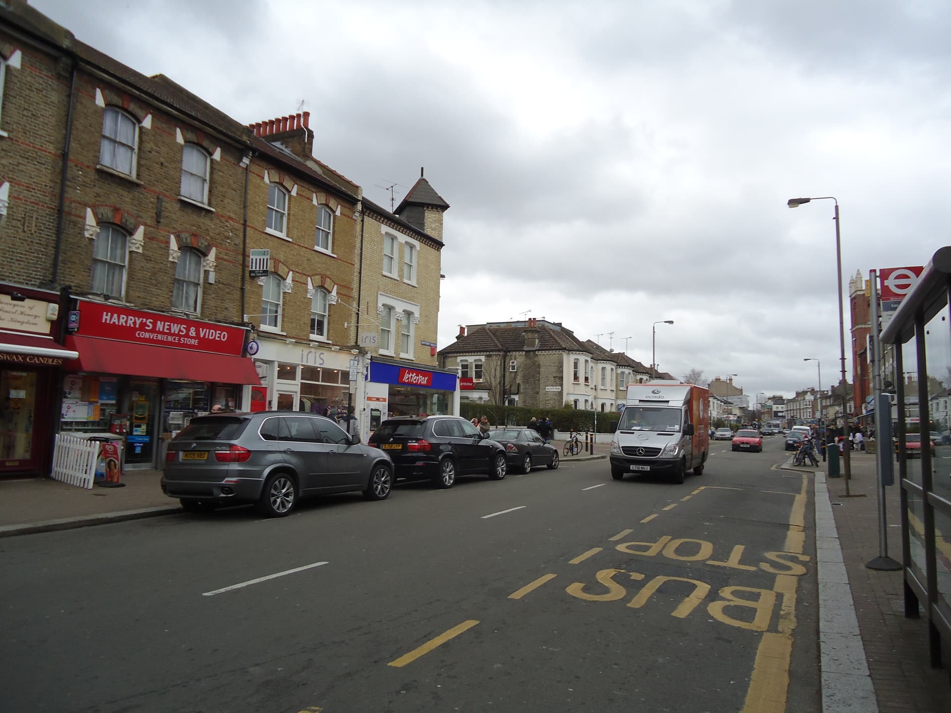 Parade of Victorian brick shops and parked cars along a London high street with a bus stop marking
