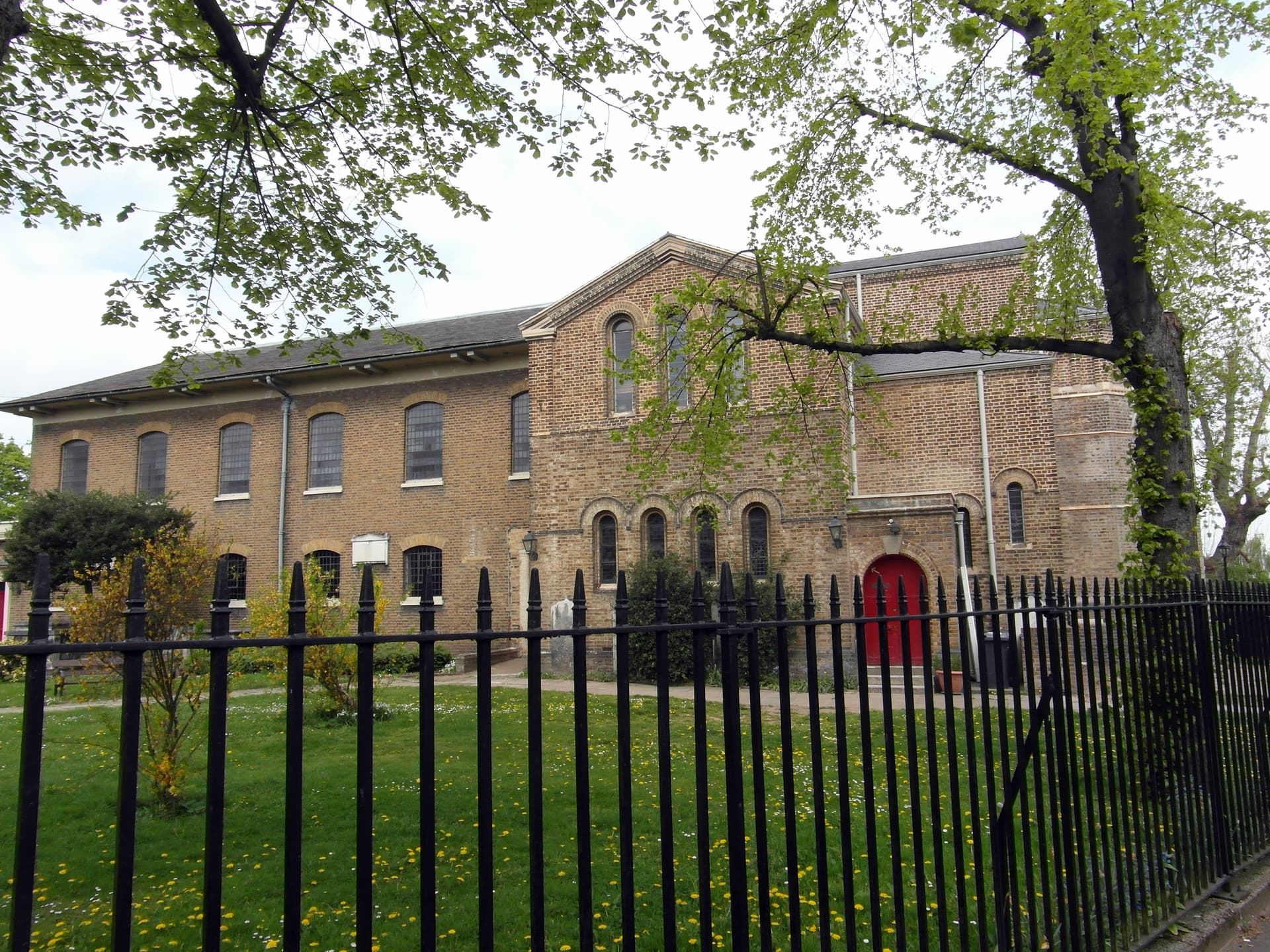 Yellow-brown brick church with arched windows and a red door, seen through black railings and spring trees