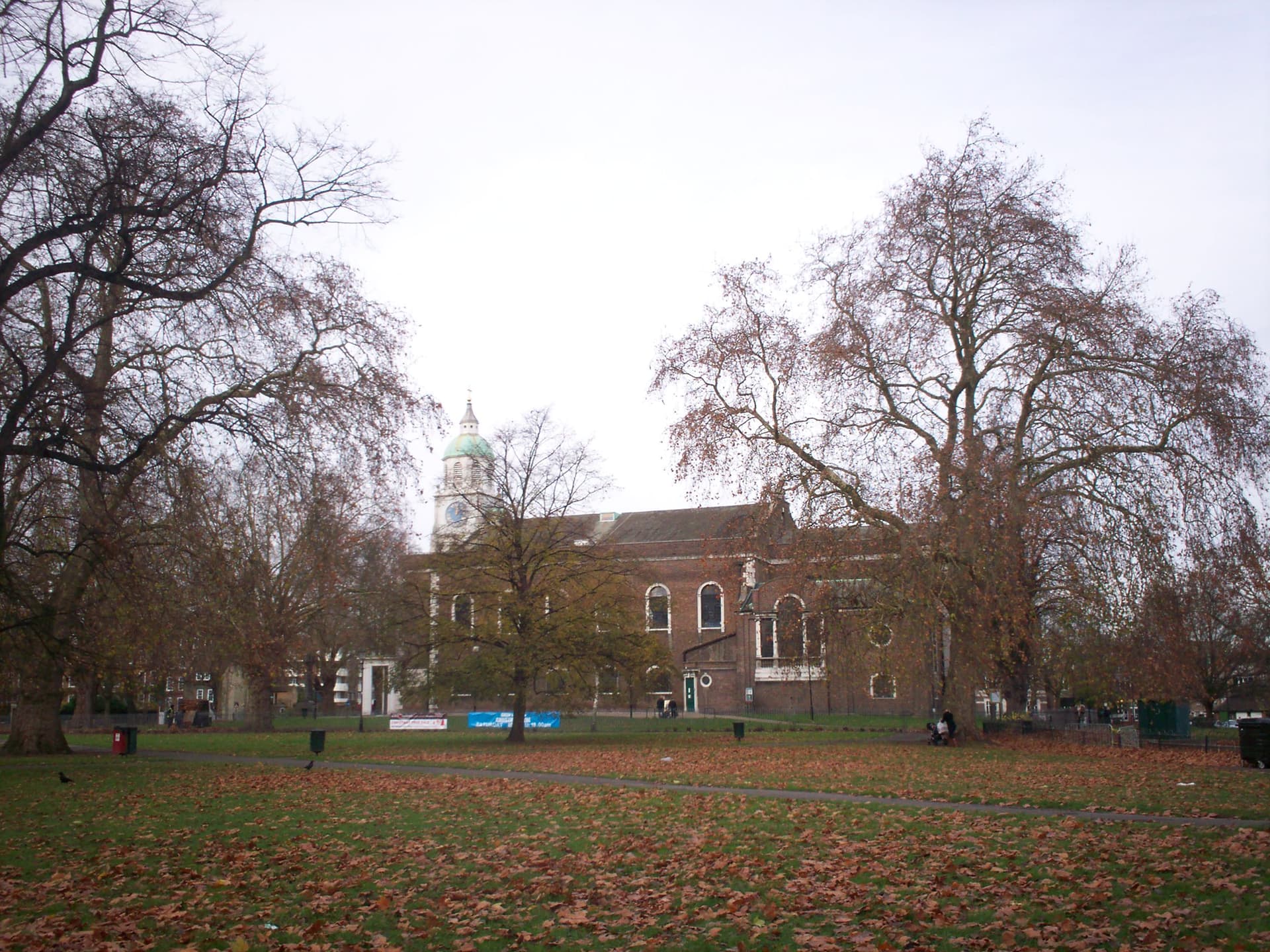 Brick church with a green-domed clock tower seen across a leaf-strewn common beneath bare autumn trees