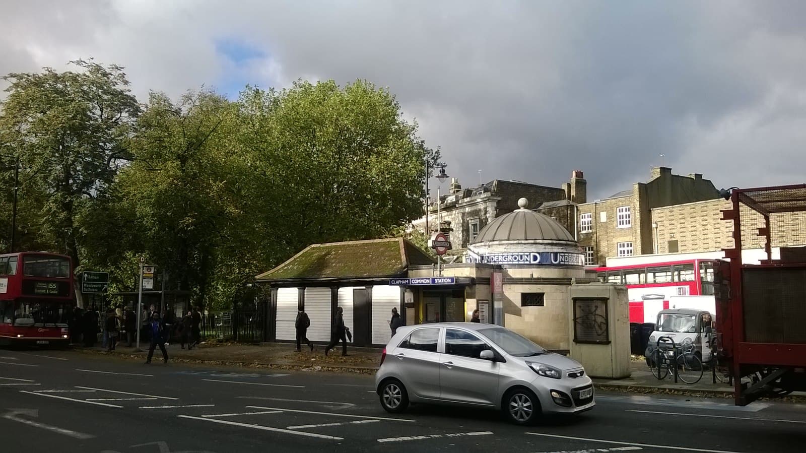 Clapham Common Underground station entrance with domed roundel building, trees, red London buses and a silver car on the road