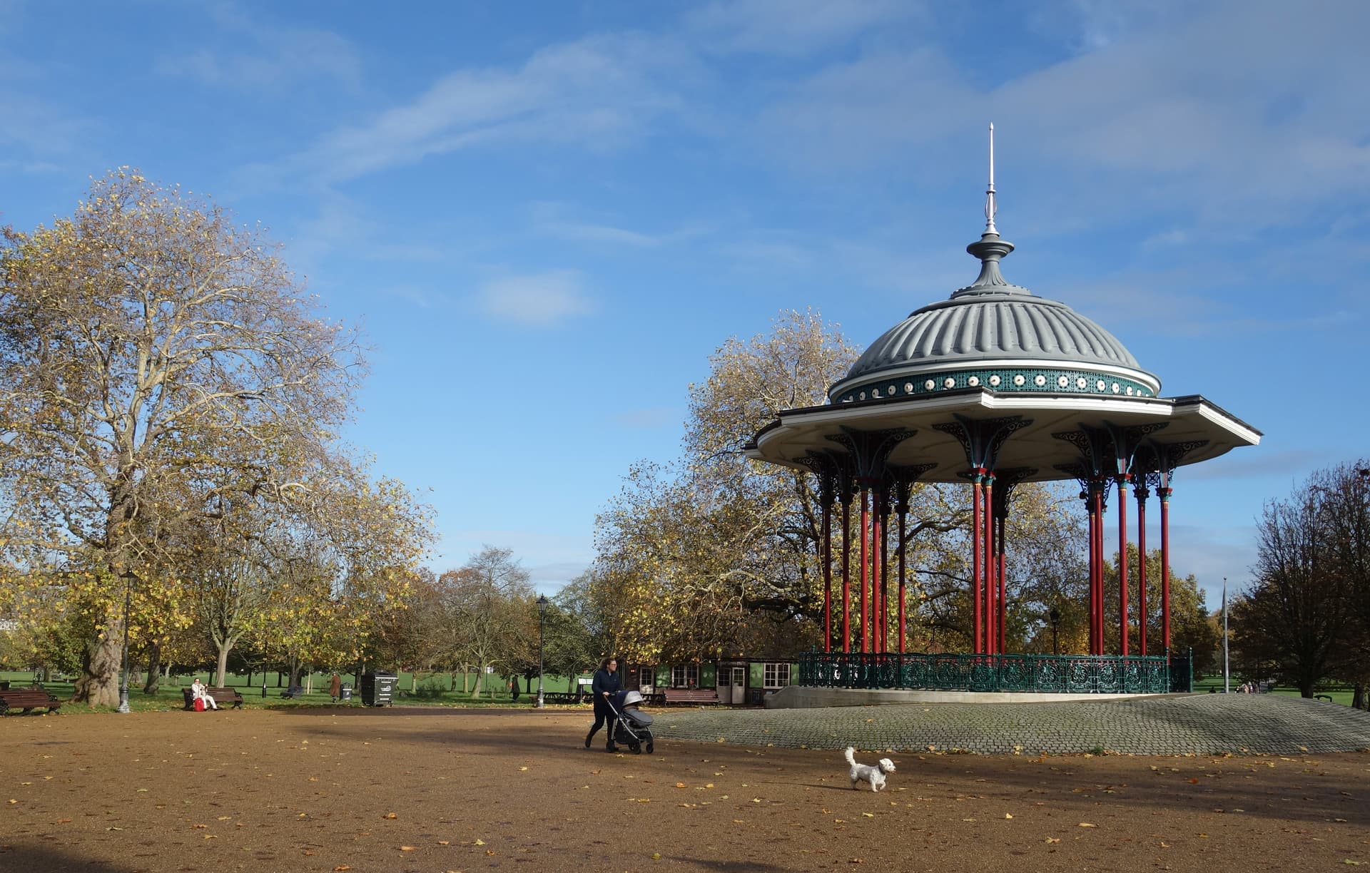 Victorian bandstand with red columns and grey dome on Clapham Common, with a woman pushing a pram and a small white dog